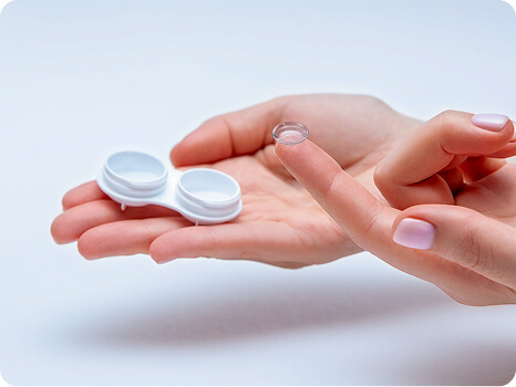 Hand holding a contact lens case while another hand places a soft contact lens on a fingertip.
